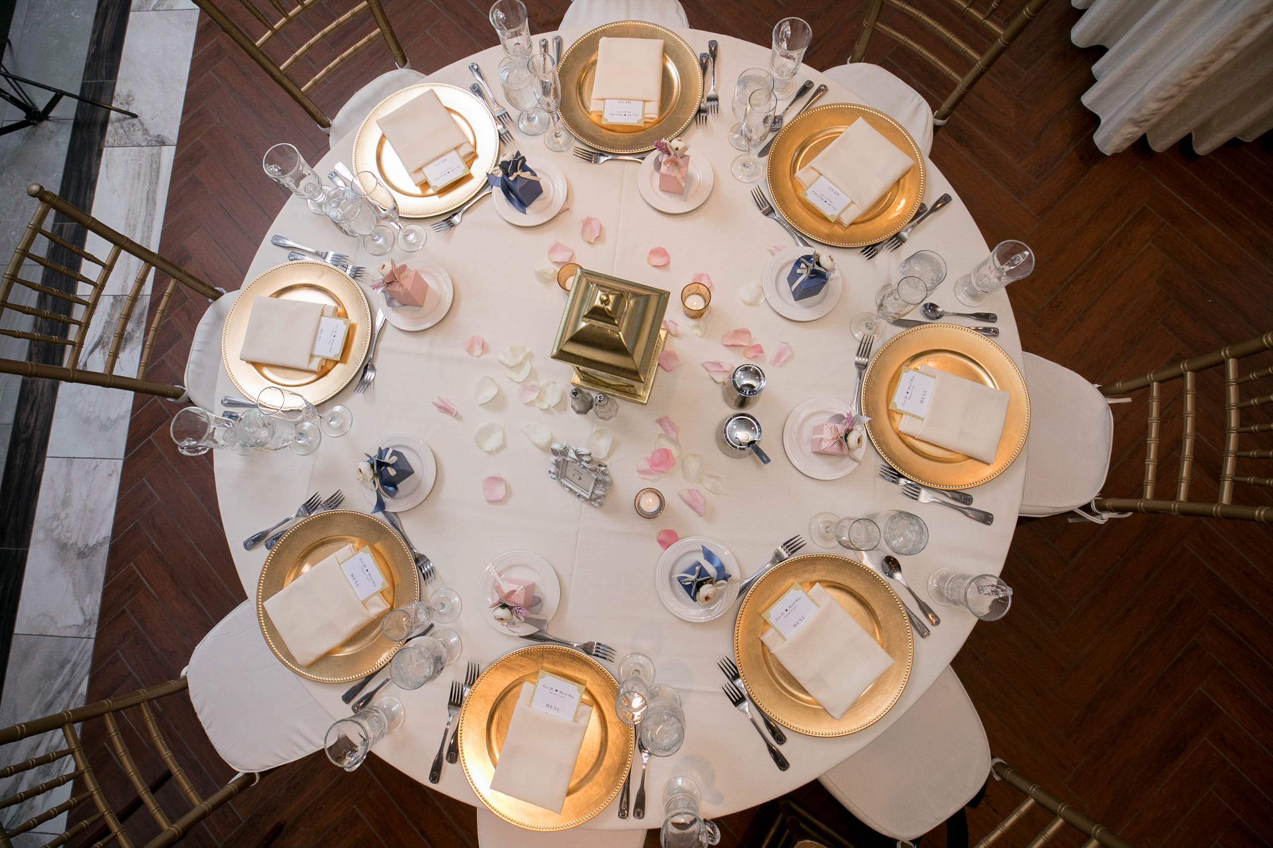 An overhead shot of a table covered with a white table cloth shows place settings, a gold lantern, and white and pink rose petals. 